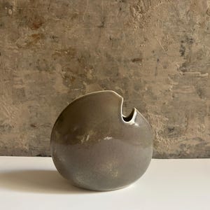 Op de afbeelding: Een ronde keramische vaas met een gladde, glanzende, taupekleurige glazuurlaag. De vaas heeft een asymmetrische, gebogen bovenrand met een kleine, duidelijke opening aan de rechterkant, wat zorgt voor een halvemaanvormig silhouet.