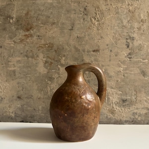 Può includere: Una piccola brocca in ceramica arrotondata con un unico manico e un beccuccio stretto. La superficie presenta una smaltatura marrone screziata e testurizzata con macchie più scure e chiare. Un piccolo motivo floreale in rilievo è visibile.
