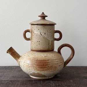 Op de afbeelding: Een keramische theepot met een gestapeld, tweedelig ontwerp met een gespikkeld beige en bruin glazuur. De basis is rond met een gebogen handvat en tuit, terwijl het bovenste gedeelte cilindrisch is met twee kleine zijhandvatten en een deksel.