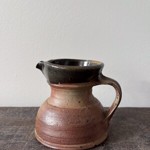 Op de afbeelding: Een kleine keramische kan met een ronde basis en een brede, uitlopende rand. Het lichaam heeft een getextureerde, aardse bruine en beige gespikkelde afwerking, terwijl de bovenrand en de binnenkant zijn geglazuurd in een glanzend antracietzwart.