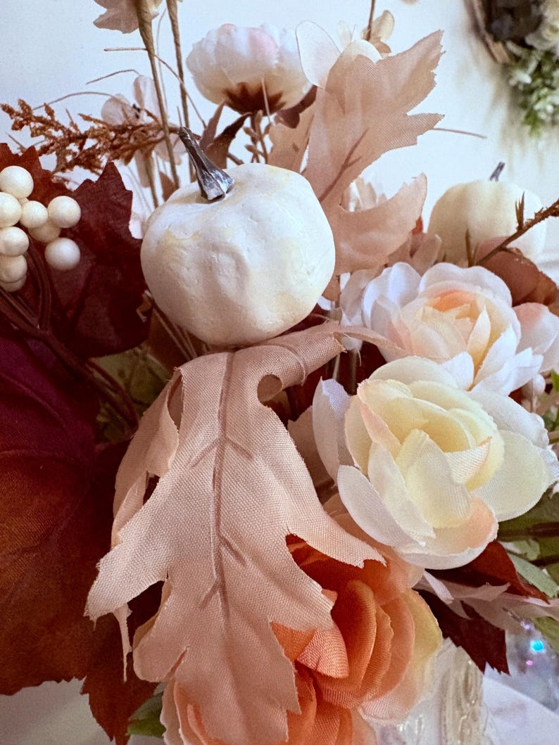 Puede incluir: Arreglo floral oto&ntilde;al con calabazas blancas, rosas color melocot&oacute;n y hojas artificiales en tonos marr&oacute;n y melocot&oacute;n. El arreglo incluye bayas blancas y diversas flores, creando un ambiente c&aacute;lido y acogedor, ideal para decoraci&oacute;n.