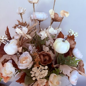 Puede incluir: Arreglo floral con flores blancas, crema y melocot&oacute;n, incluyendo rosas y peon&iacute;as, con acentos de hojas marrones y peque&ntilde;as calabazas blancas. Las flores est&aacute;n dispuestas en un jarr&oacute;n blanco, creando un centro de mesa oto&ntilde;al.