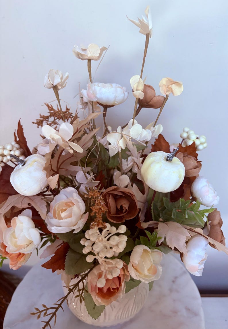 Puede incluir: Arreglo floral con flores blancas, crema y melocot&oacute;n, incluyendo rosas y peon&iacute;as, con acentos de hojas marrones y peque&ntilde;as calabazas blancas. Las flores est&aacute;n dispuestas en un jarr&oacute;n blanco, creando un centro de mesa oto&ntilde;al.