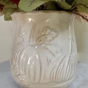 Puede incluir: Jarr&oacute;n de cer&aacute;mica color crema con dise&ntilde;os en relieve de calabazas y hojas. El jarr&oacute;n tiene un borde ligeramente acampanado y una superficie texturizada. Est&aacute; lleno de flores y follaje artificiales de oto&ntilde;o.