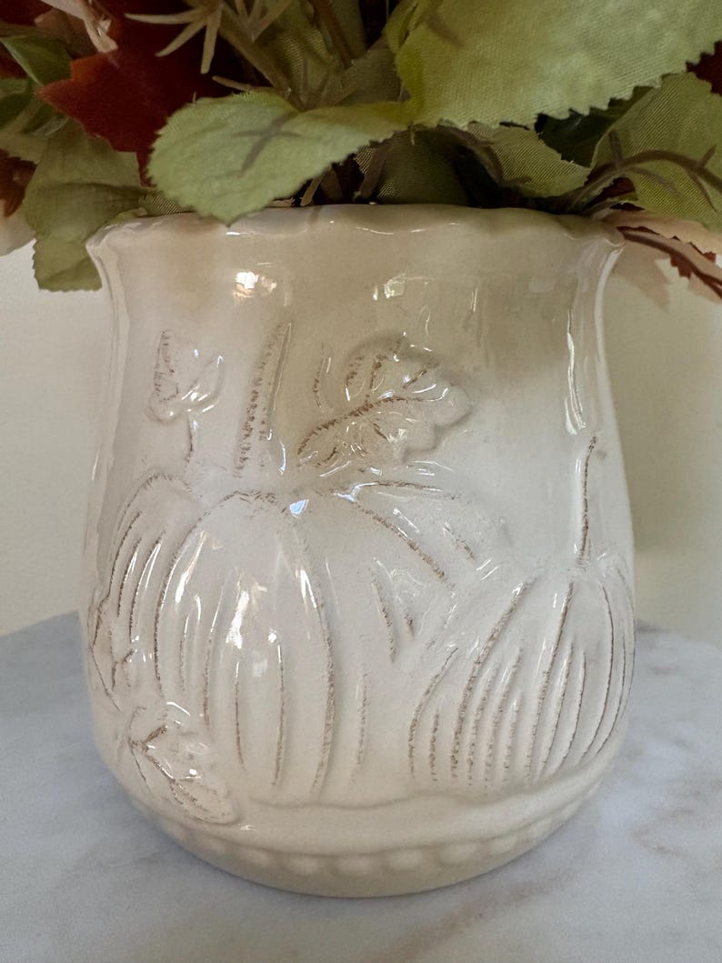 Puede incluir: Jarr&oacute;n de cer&aacute;mica color crema con dise&ntilde;os en relieve de calabazas y hojas. El jarr&oacute;n tiene un borde ligeramente acampanado y una superficie texturizada. Est&aacute; lleno de flores y follaje artificiales de oto&ntilde;o.