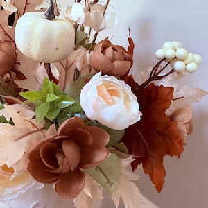 Puede incluir: Un arreglo floral con tem&aacute;tica oto&ntilde;al que presenta una peque&ntilde;a calabaza blanca, rosas color crema y varias hojas marrones y beige. El arreglo incluye bayas blancas y follaje verde, creando una est&eacute;tica c&aacute;lida y oto&ntilde;al. Las flores est&aacute;n en un jarr&oacute;n blanco.