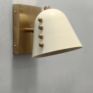 May include: A wall-mounted light fitting featuring a brass base and a cream-coloured, bell-shaped shade. The shade has four brass rivets. The fitting is mounted on a light grey wall.