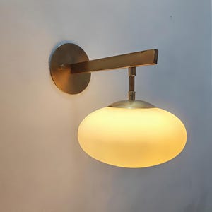 May include: A wall-mounted light fixture with a brass finish. It features a round backplate, a rectangular arm, and a frosted, oval-shaped glass shade. The light is emitting a warm, yellow glow.