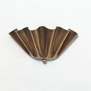May include: A brass-colored wall sconce with a scalloped, fan-like design. The sconce has a small, round detail at the bottom. The fixture is mounted on a white wall.