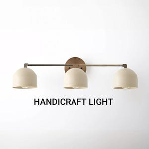 May include: A three-light fitting with a bronze finish. The light shades are cream-coloured and dome-shaped. The text "HANDICRAFT LIGHT" is printed below the fitting.