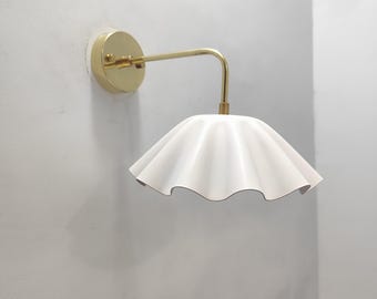 May include: A wall-mounted light fixture with a gold-colored arm and a white, ruffled lampshade. The fixture is attached to a circular gold base. The lampshade has a wavy, scalloped edge.