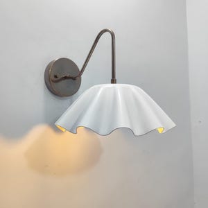 Op de afbeelding: Een witte, gerimpelde lampenkap hangt aan een bronzen arm en een cirkelvormige wandbevestiging. De binnenkant van de kap gloeit met een warm licht. De armatuur is op een witte muur gemonteerd, wat een eenvoudig, elegant ontwerp creëert.