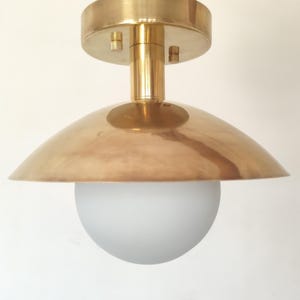 May include: A brass-colored ceiling light fixture with a rounded shade and a frosted glass globe. The fixture has a cylindrical stem and a circular base. The light is suspended from a thin wire.