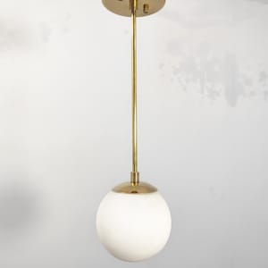 May include: A modern pendant light fixture with a brass finish. The fixture features a white globe shade and a brass rod extending from a circular ceiling mount. The overall design is minimalist and elegant, suitable for various interior styles.