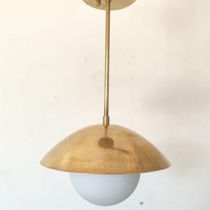 May include: A brass pendant light fixture with a rounded shade and a white globe. The fixture has a circular ceiling mount and a long, straight rod. The shade is a warm gold color, and the globe is a soft white.