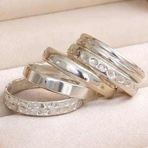 May include: A collection of silver rings displayed inside a beige jewelry box. The rings vary in design, including smooth bands and those with textured or gem-like details. The box is embossed with the text "KO LUXURY Jewelry".