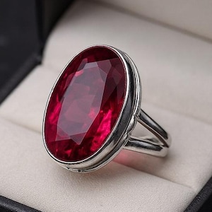 May include: A silver ring featuring a large, oval-cut, deep red gemstone, displayed in an open black jewellery box with a white interior. The ring has a split band design. The box is set against a dark background.