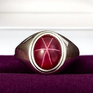 Może przedstawiać: Srebrny pierścionek z owalnym rubinem w kształcie gwiazdy. Pierścionek jest prezentowany w czarnym pudełku z białym wnętrzem i fioletową aksamitną poduszką. Rubin ma sześcioramienny wzór gwiazdy.
