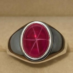 May include: A silver ring with an oval, star-shaped ruby gemstone. The ruby is a deep red color, and the ring is set in a polished silver band. The ring is displayed in a beige jewelry box.