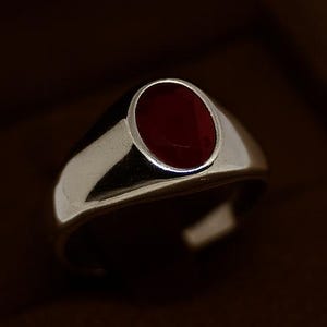 May include: A silver ring with a polished finish, featuring a deep red, oval-shaped stone. The ring is set in a classic bezel setting, presented in a brown jewellery box. The ring's design is simple and elegant.