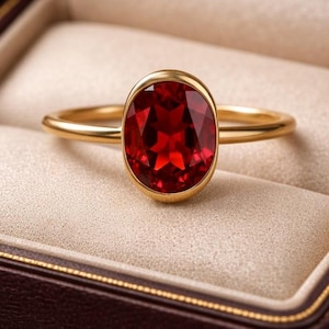 May include: A gold ring featuring a large, oval-cut, red gemstone. The ring is displayed in an open, brown jewellery box with a cream interior. The box highlights the ring's setting and the gemstone's facets.