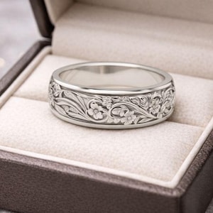 K&ouml;nnte beinhalten: Ein silberner Ring mit aufwendigem Blumendesign, pr&auml;sentiert in einer braunen Ringschatulle mit cremefarbenem Innenraum. Der Ring weist detaillierte Schnitzereien von Blumen und Bl&auml;ttern auf, die einen klassischen und eleganten Stil zeigen.