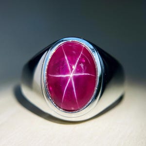 Puede incluir: Un anillo de plata con una gran gema de rubí ovalada. El rubí muestra un efecto de estrella de seis rayos, con un tono rojo intenso y una superficie pulida. El anillo tiene una banda de plata pulida simple.