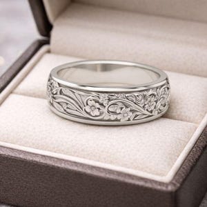 K&ouml;nnte beinhalten: Ein silberner Ring mit aufwendigem Blumendesign, pr&auml;sentiert in einer braunen Ringschatulle mit cremefarbenem Innenraum. Der Ring weist detaillierte Schnitzereien von Blumen und Bl&auml;ttern auf, die einen klassischen und eleganten Stil zeigen.