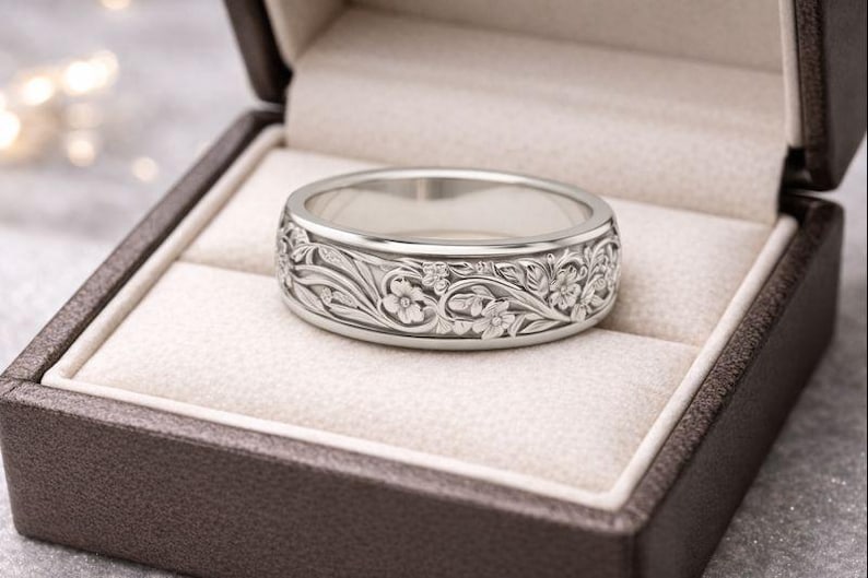 K&ouml;nnte beinhalten: Ein silberner Ring mit aufwendigem Blumendesign, pr&auml;sentiert in einer braunen Ringschatulle mit cremefarbenem Innenraum. Der Ring weist detaillierte Schnitzereien von Blumen und Bl&auml;ttern auf, die einen klassischen und eleganten Stil zeigen.