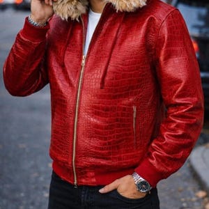 Könnte beinhalten: Rote Lederjacke mit Kapuze mit Kunstfellfutter. Die Jacke hat ein strukturiertes, krokodilartiges Muster, einen goldenen Reißverschluss und rote gerippte Bündchen und Saum. Die Person trägt ein weißes Hemd und eine schwarze Jeans.