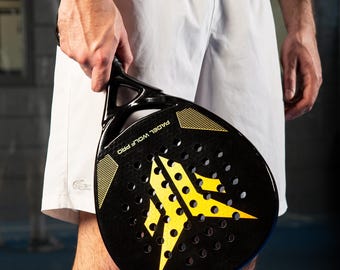 May include: Black and yellow padel racket with the text "PADEL WOLF PRO". The racket has a perforated surface and a yellow starburst design. The person is wearing white athletic shorts and a long-sleeve shirt.
