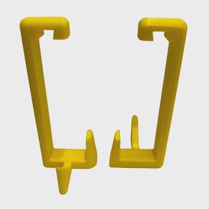 May include: Two yellow plastic clips, each with a rectangular top and a long, angled arm. One clip has a small, vertical post. The clips are designed to hold or secure items. The background is a solid, neutral color.