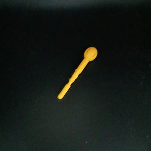Puede incluir: Una cuchara de plástico amarilla con una cabeza redondeada y un mango largo y delgado. La cuchara está en ángulo diagonal sobre un fondo negro. La cuchara parece ser una sola pieza de plástico moldeado.