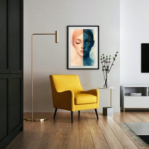 Może przedstawiać: Oprawiony wydruk przedstawiający podzieloną twarz, jedną stronę w odcieniach brzoskwini, drugą w głębokim niebieskim. Żółty fotel z czarnymi nogami znajduje się pod grafiką. Złota lampa podłogowa i biały stolik boczny z wazonem kwiatów dopełniają scenę.