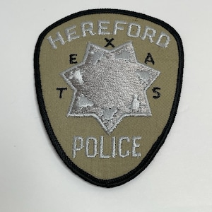 May include: A shield-shaped embroidered patch with a tan background and a black border. It features the silver text HEREFORD at the top and POLICE at the bottom. A silver seven-pointed star is in the center with the black letters T, E, X, A, S.