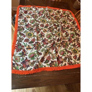 May include: A square fabric cloth featuring a white background with a dense, multi-colored floral pattern in shades of red, green, yellow, and brown. The edges are finished with a bright orange fringe trim that borders all four sides.