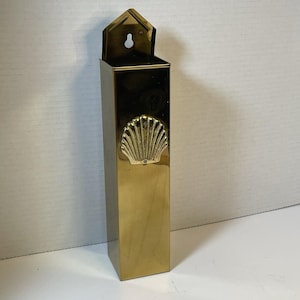 Könnte beinhalten: Ein hoher, rechteckiger Wandbehälter aus poliertem Messing mit einer spitzen Oberseite und einem Schlüssellochschlitz zur Befestigung. Die Vorderseite ist mit einem mittig platzierten, geprägten Muschelmotiv verziert und hat eine glänzende Oberfläche.