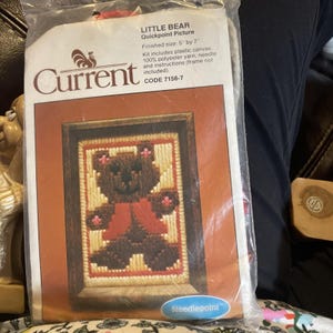 May include: A Current brand Little Bear Quickpoint Picture kit in a clear plastic bag. The packaging features a brown bear graphic with a red vest, finished size 5 inches by 7 inches, including plastic canvas, 100% polyester yarn, and a needle.