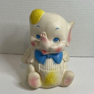 Peut inclure: Une figurine d'éléphant en caoutchouc blanc portant une casquette jaune, un nœud papillon bleu et une salopette blanche avec un emblème de lapin jaune sur le devant. L'éléphant a des joues roses, des yeux bleus et des accents roses sur les oreilles.