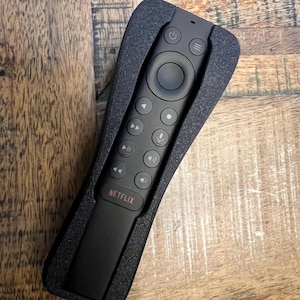 May include: A black remote control with multiple buttons, including a power button, volume controls, and a microphone button. The word "NETFLIX" is printed in red at the bottom. The remote is in a black foam holder.