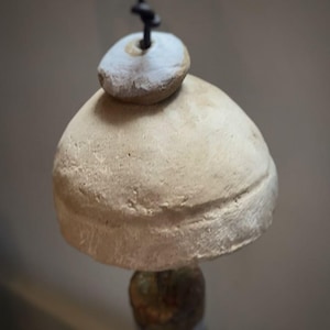 May include: A handmade wind chime with a natural, earthy appearance. The chime has a large, beige, bell-shaped body topped with a smaller, round, white-painted stone. A black cord suspends the chime.