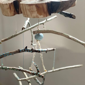 May include: A handcrafted mobile featuring natural wood slices, small acorn-like beads, and branches. The branches are adorned with blue and green accents. The mobile is suspended by thin strings, creating a delicate and organic aesthetic.