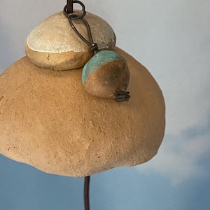 May include: A handmade wind chime with natural stone components. It features a large, rounded, light brown stone base, a smaller stone on top, and a small, teal and brown accent piece. The design is completed with a leather cord and wire.