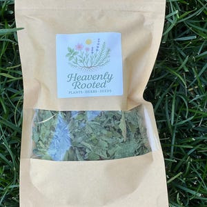 May include: A brown paper bag with a clear window, filled with dried green herbs. The bag has a white label with the text "Heavenly Rooted" and a floral design. The bag is sitting on green grass.
