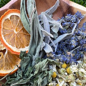 May include: A wooden bowl filled with dried herbs and citrus. The arrangement includes dried orange slices, sage, lavender, chamomile, and other green herbs. The herbs are various shades of green, blue, and yellow.