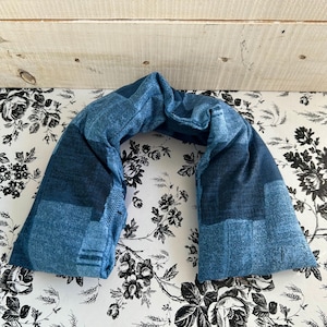 May include: A blue, U-shaped neck wrap with a patchwork design. The wrap is made of a soft, textured fabric and is designed to be worn around the neck and shoulders. The background is a floral pattern.