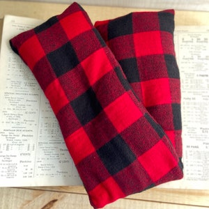 May include: Two rectangular, red and black buffalo plaid fabric items. The items are likely filled with a material to provide warmth or support. The fabric has a classic, rustic pattern, with large, evenly spaced squares. The items are resting on a vintage paper background.