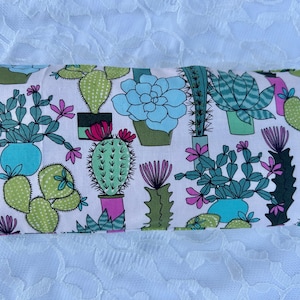 May include: Rectangular fabric item with a repeating pattern of various cacti and succulents in pots. The plants are in shades of green, blue, and pink, set against a white background. The item is likely a pillow or a similar home decor item.
