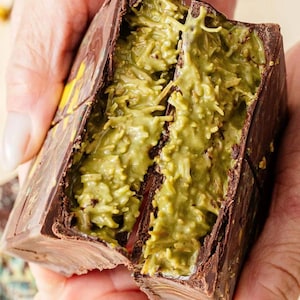 May include: A chocolate confection, broken open to reveal a green filling. The dark chocolate exterior encases a textured, light green interior. The dessert is held in a person's hands, showcasing the contrasting colors and textures.