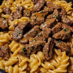 Può includere: Primo piano di un piatto di pasta con fusilli e pezzi di carne in salsa cremosa. La carne è marrone e sembra essere condita con erbe. La pasta è ricoperta da una salsa chiara, servita in una ciotola scura.